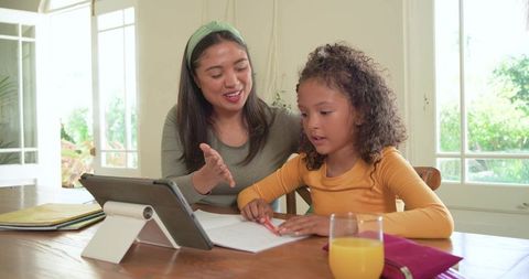 Woman guiding child with tablet during home learning session at sunlit dining table