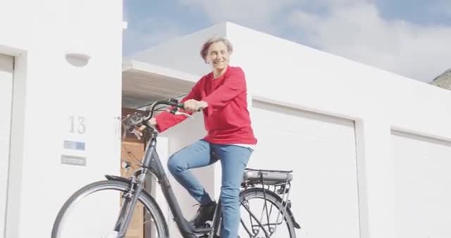 Senior Caucasian woman with short grey hair riding a bicycle, radiating joy and energy while enjoying an outdoor activity on a sunny day. Her bright red sweater adds vibrancy to the scene. This visual can be used for promoting healthy lifestyles, retirement community advertisements, or inspiring stories about active living in older age.