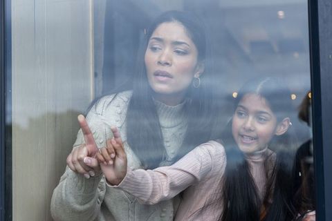 Indian mother and daughter looking through window pointing outside wearing cozy sweaters