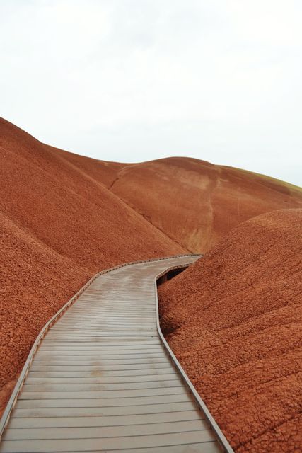 The image depicts a solitary wooden pathway cutting through rugged, undulating red hills. This dramatic arid landscape highlights the dry, rich red earth and sparse vegetation, creating a striking contrast against the wooden pathway. Ideal for use in travel brochures, hiking guides, geology topics, or articles focusing on natural beauty and remote destinations.