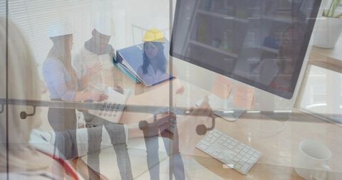 Cross-disciplinary team discussing project at desk with monitor, binder, yellow hard hat