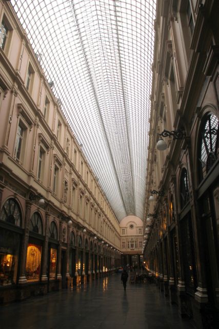 Depicts a historic indoor shopping arcade with an elegant glass ceiling, creating a luxurious and inviting atmosphere. Features ornate designs and rows of retail stores. Ideal for use in articles about historic architecture, luxury shopping experiences, European travel destinations, or architectural design. Also suitable for use in flyers, brochures, and websites promoting shopping centers or architectural tourism.