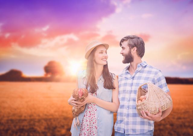 This image shows a happy couple standing in a rural field during sunset. One person holds a basket of fresh eggs, while the other holds a chicken. The setting sun creates a warm, tranquil atmosphere. Perfect for use in advertisements, blogs, or articles related to farming, sustainable living, rural lifestyles, agriculture, or romantic getaways.
