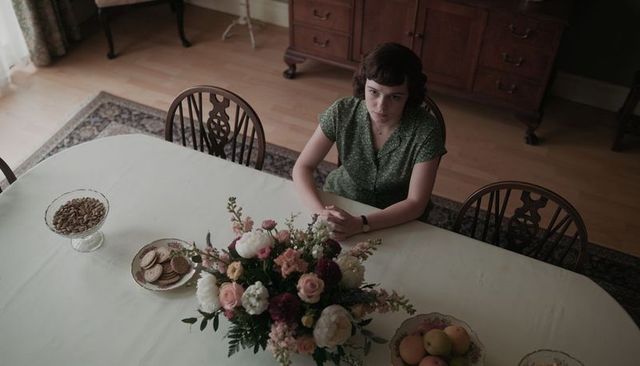 Woman sitting at dining table clasping hands beside floral centerpiece in vintage home