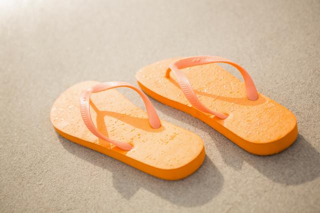 Bright orange flip-flops placed on sandy beach, perfect for summer vacation and beach-themed content. Useful for travel blogs, summer clothing advertisements, and relaxation or leisure articles.
