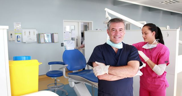 Dentist and assistant preparing for a dental procedure in a modern clinic. Dentist wearing a navy blue scrubs, gloves, and a facial mask, and the assistant in pink scrubs adjusting her gloves. Ideal for health care, professional services, dental care advertising, clinic brochures, or educational materials on dental hygiene and teamwork in medical settings.