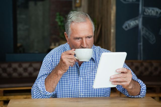 Senior man enjoying coffee while using a digital tablet in a cozy cafe. Ideal for illustrating modern technology use among elderly, leisure activities, and relaxed lifestyles. Suitable for articles on senior living, technology adoption, and coffee culture.