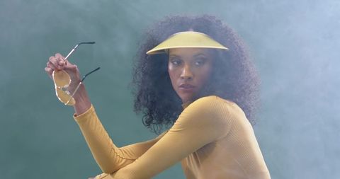 African american model posing in mustard visor and ribbed top holding sunglasses amid haze