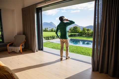 Man talking on smartphone by sliding glass door overlooking pool, lawn and mountain view