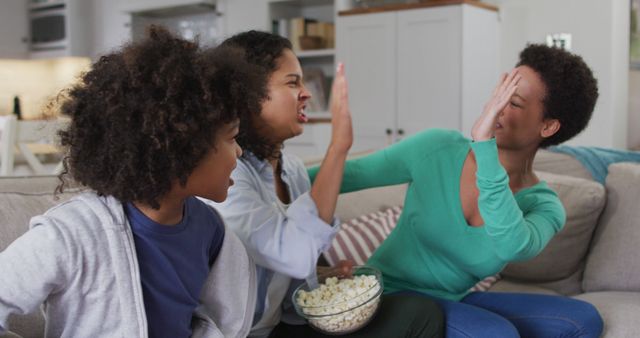 African American family spending quality time in the living room, enjoying a movie night together. Two family members give high-fives while smiling and sitting on the couch with a child who is also smiling. The family shares a bowl of popcorn. Perfect for advertisements promoting family time, snacks, or lifestyle content focused on familial bonding and entertainment.