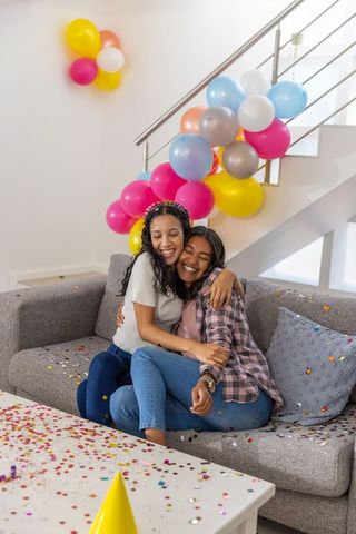 Diverse friends hugging on sofa celebrating birthday with balloons and confetti