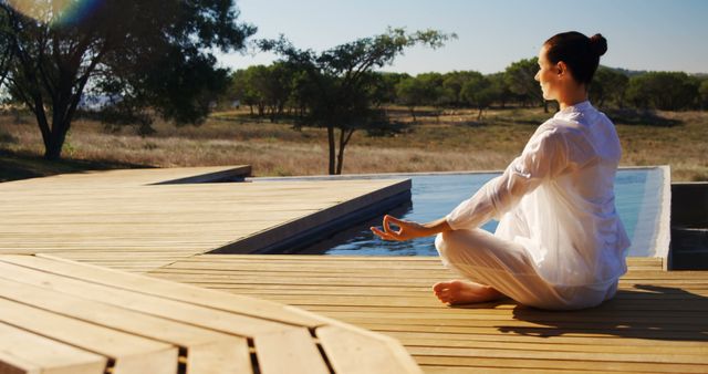 Woman meditating outdoors on wooden deck by infinity pool during sunrise. She is sitting in lotus position wearing a light, flowing outfit. Surroundings are natural with greenery and clear sky, evoking serenity and tranquility. Ideal for use in wellness advertisements, mindfulness or yoga practices, travel brochures, and relaxation or meditation guides.