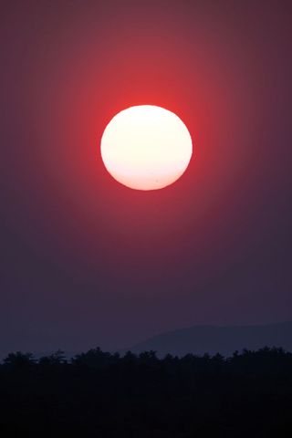 Sun glowing in deep red sky over forest silhouette and rolling hills at tranquil dusk