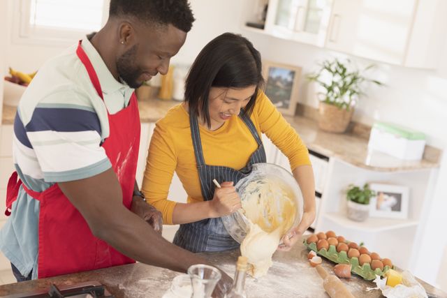 A cheerful couple enjoys baking in a bright, modern kitchen. This image is ideal for illustrating concepts of home life, family bonding, and multicultural relationships. Perfect for use in lifestyle blogs, domestic product advertisements, and social media posts promoting togetherness and home activities.