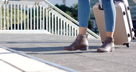 Traveler walking with rolling suitcase on urban walkway in jeans and ankle boots