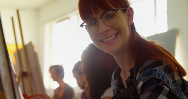 A young woman with red hair wearing glasses is smiling and holding a paintbrush while painting on a canvas in an art class. Other students are seen painting in the background, creating a collaborative and creative atmosphere. This image is ideal for illustrating concepts related to education, arts, creativity, teamwork, and artistic workshops.