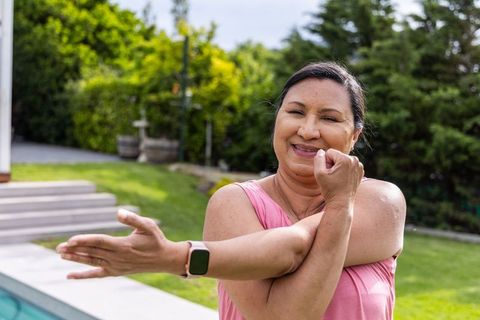 Mature woman doing shoulder stretch poolside in backyard wearing pink tank and smartwatch