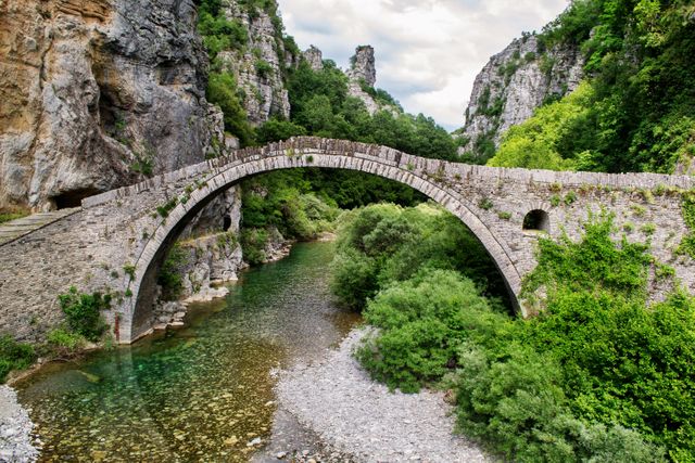 Depicting a historic stone bridge arching over a pristine river in a tranquil mountainous landscape, this image showcases ancient architecture amidst lush greenery. Ideal for use in travel brochures, heritage site promotions, and environmental conservation materials.