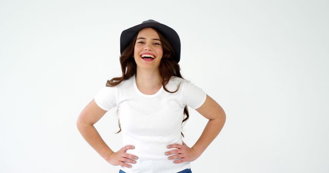 This stock photo features a cheerful young woman with long hair wearing a hat and a white shirt, smiling confidently and posing with hands on hips. Perfect for promotions, marketing materials, lifestyle blogs, and advertising campaigns that need a positive and happy representation.