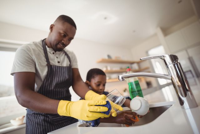 Father and son washing dishes together in a bright, modern kitchen. Both wearing rubber gloves, the pair engages in an everyday household chore, promoting family bonding and teamwork. Suitable for use in articles or advertisements about family life, domestic responsibilities, or teamwork.