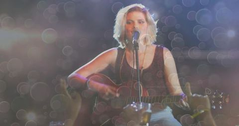 Teen female singer-songwriter strumming acoustic guitar onstage under dreamy bokeh lights