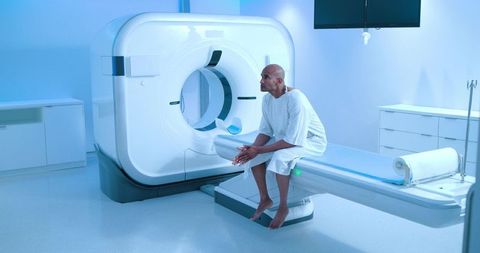 Patient sitting on ct scanner table wearing hospital gown, clasping hands in clinical imaging room