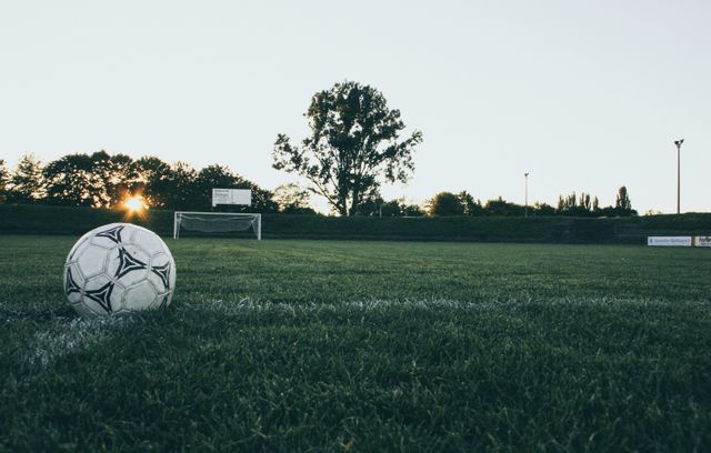 Soccer ball resting on a green football field during sunset, with a goal post and greenery in the background. This serene setting is perfect for inspiring sports-themed designs, training session promotions, or outdoor activity advertisements.