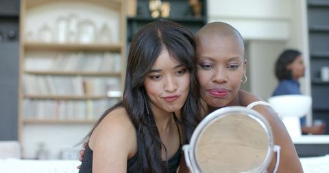 Leaning friends puckering while checking makeup in round mirror on cozy sofa