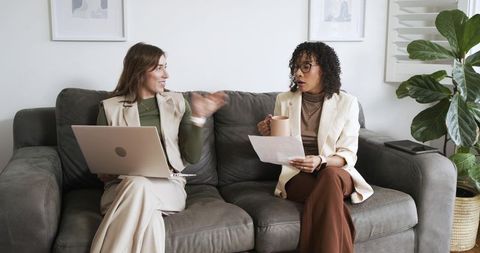 Mid-adult women collaborating on laptop and document on gray sofa discussing project in home office