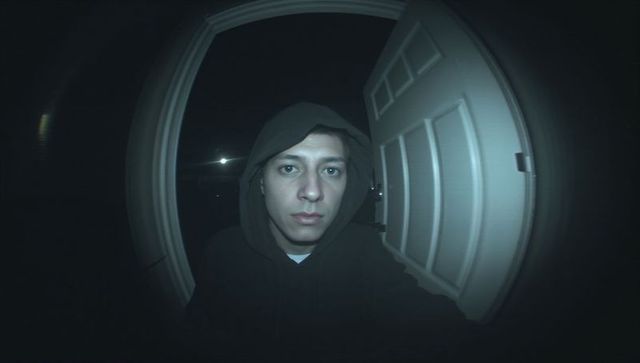 Hooded man looking into doorbell camera at front porch nighttime fisheye infrared close-up