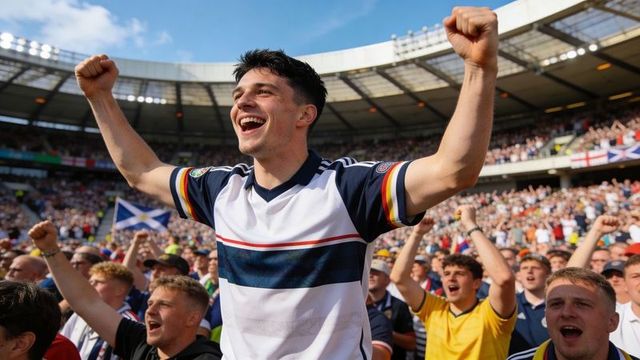 Young male fan cheering and raising arms in packed stadium crowd with saltire flag
