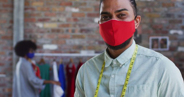 Fashion designer wearing red face mask and measuring tape stands in studio with colleague in background examining garments. Suitable for use in articles related to fashion design, tailoring business, health and safety measures in creative fields.