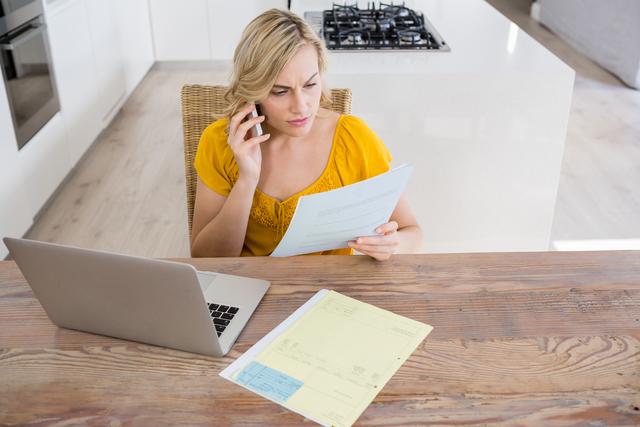 Young woman sitting at kitchen table, talking on mobile phone while reviewing bills. Laptop and documents spread on wooden table. Ideal for illustrating financial planning, home budgeting, or modern communication.