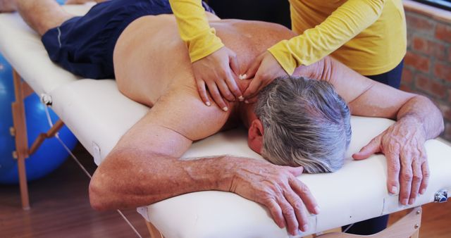 Elderly man receiving shoulders and back massage from a physiotherapist on a treatment table in a clinic. Professional massage therapy promoting relaxation and pain relief for senior patients. Ideal for healthcare advertisements, geriatric wellness campaigns, and physical therapy educational materials.