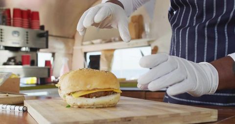 Gloved cook assembling seeded cheeseburger on wooden board in cafe with espresso machine