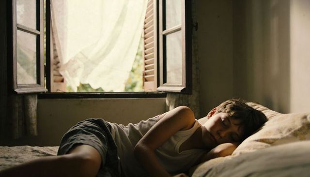 Sleeping teenage boy resting on bed by open window with sunlit curtains and breeze