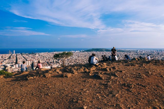 Group of tourists sitting on a mountain peak with a panoramic view of the city and the ocean on a sunny day. Ideal for promoting travel destinations, showcasing landscape photography, or illustrating adventures in nature. Suitable for websites related to tourism, travel blogs, and adventure activities.