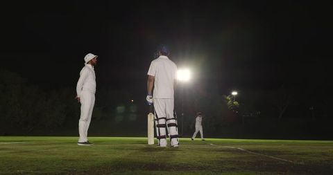 Backlit night cricket under floodlights with indian umpire standing and batsman at crease