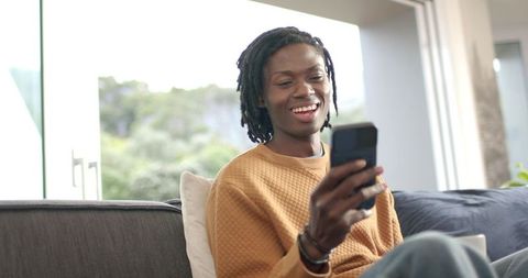 African American man smiling with smartphone on couch by window in mustard sweater