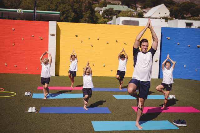 Coach leading a yoga session with school kids in a schoolyard. Children are following the coach's lead, practicing balance and stretching on yoga mats. Ideal for use in educational materials, fitness and health promotions, and community activity advertisements.