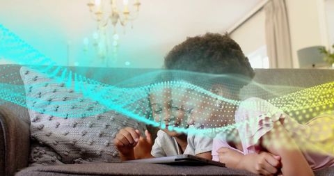 Two siblings lying on sofa watching tablet with colorful dotted data wave overlay, cozy family tech