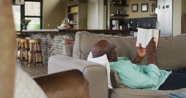 African american senior man relaxing, lying on couch reading book. retirement lifestyle, spending time alone at home.