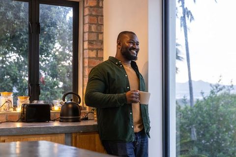 African american man gazing out window holding mug in cozy kitchen with warm candlelight