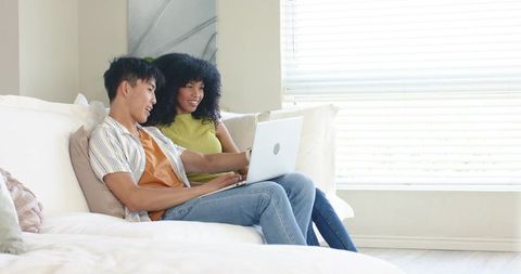 Mixed-race couple relaxing on white sofa using laptop in bright minimalist living room