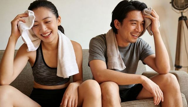 Couple wiping foreheads and laughing while cooling down on sofa after home workout
