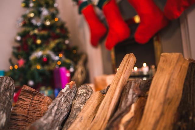 Close-up of firewood logs in front of a decorated fireplace with Christmas stockings and a tree in the background. Ideal for holiday-themed promotions, cozy winter home decor, and festive celebration advertisements.