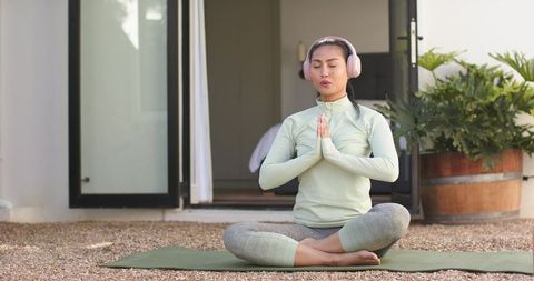 Asian woman meditating on porch in activewear with headphones practicing mindfulness