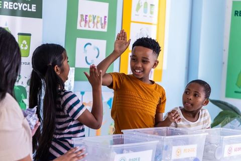 Multicultural children high-fiving and sorting recyclables at classroom recycling station