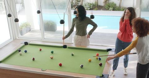 Diverse women playing pool by glass doors overlooking backyard pool, casual home hangout
