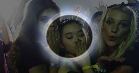 Young women blowing kisses under ring light at nightclub party with colorful haze, crowd